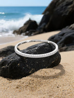 Own Your Bloom moissanite bangle bracelet placed on sand and rock with gentle sunlight reflections, elegant summer jewelry, Dallas.