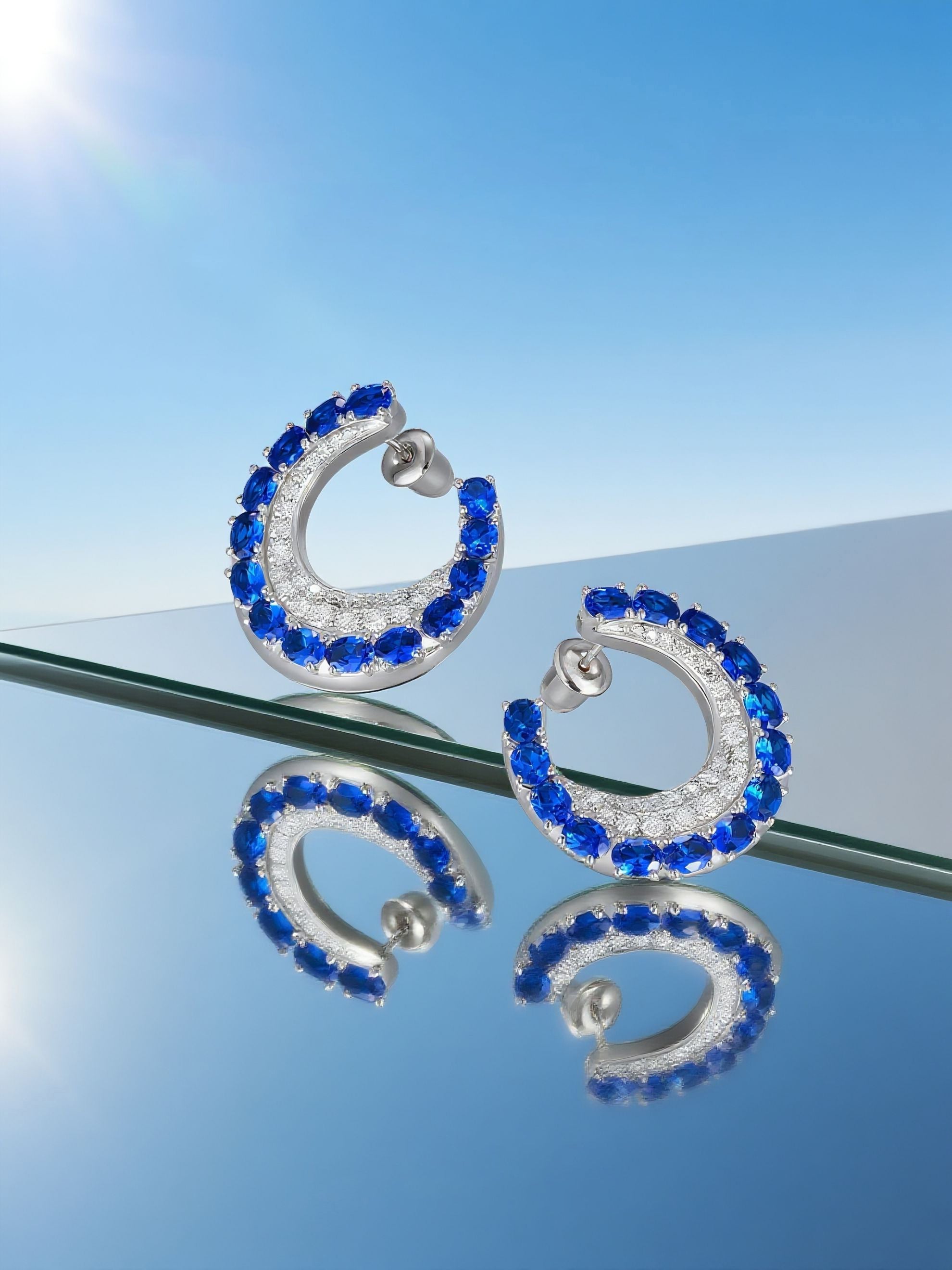 Own Your Bloom sapphire hoop earrings displayed on mirrored surface under bright sunlight, capturing gemstone reflections, Miami.