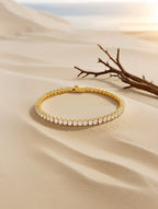 Own Your Bloom Radiant Rhythm Gold Tennis Bracelet displayed on fine sand with sunlight reflections, coastal luxury, Miami.
